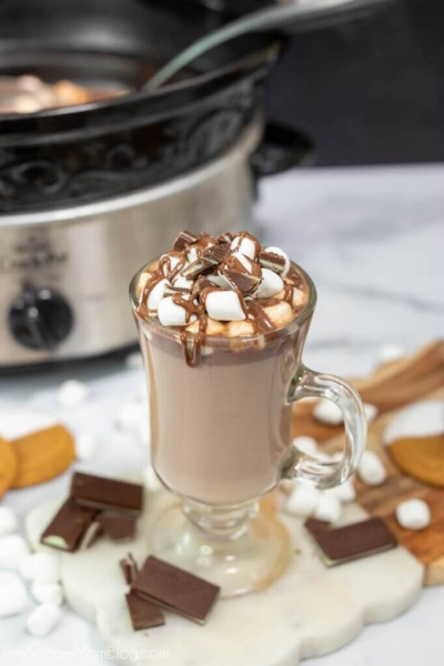 Hot chocolate in glass with marshmallows and chocolate sauce, slow cooker in background and chocolate piece on the table