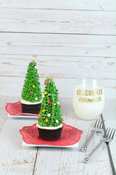 2 Cupcakes on white plates with red doilies. The cupcakes have cones on top and green icing to make them look like Christmas trees. Glass of milk and 2 forks on table