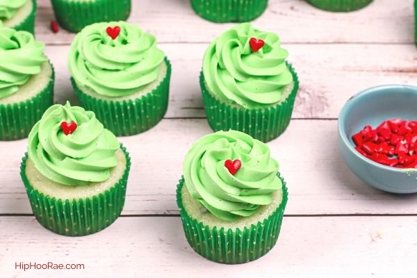 5 Grinch cupcakes on a table all have green icing and a red sugar heart for decorations