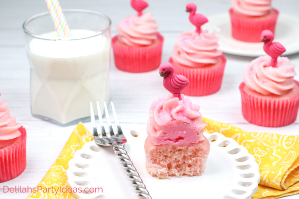 white plate with a flamingo strawberry cupcake on it, silver fork, glass of milk with straw and 3 flamingo cupcakes in background